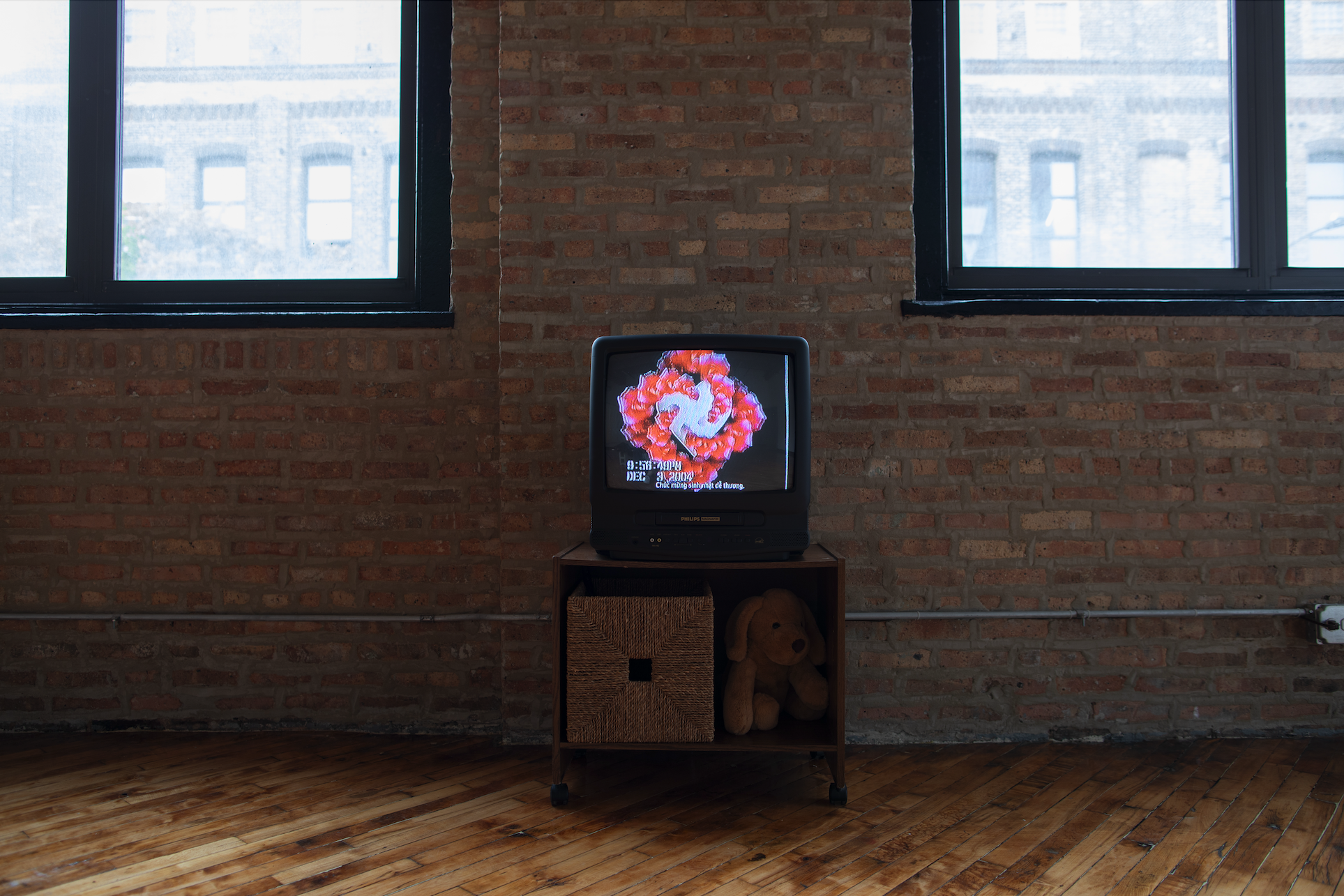 Image: In the gallery with wooden floors and exposed brick walls, a television sits between two windows showing the video work "Dear Grandpa" by Thương Hoài Trần. Photo by Khalid Ibrahim.